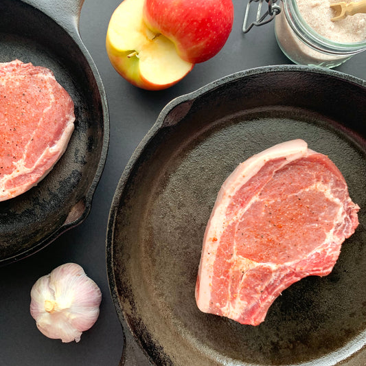 two raw pork chops each in their own pan, styled beside garlic, apple, and salt