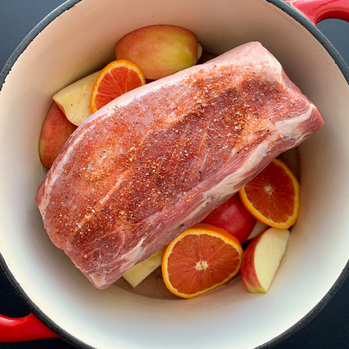 a raw pork shoulder roast, styled in a red cast iron pot with apple and orange slices, on a dark background