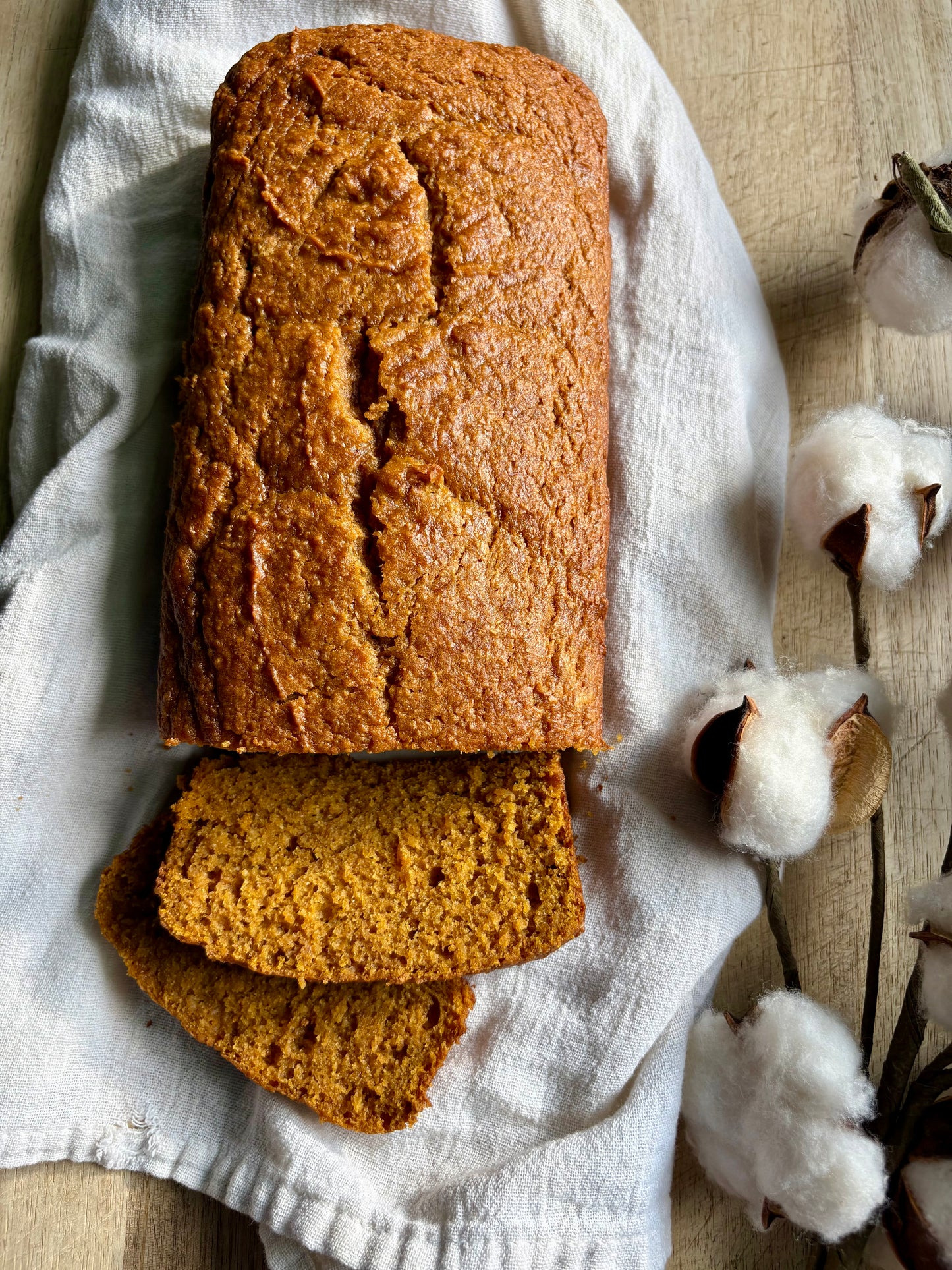 A loaf of golden-brown pumpkin bread with two slices cut and placed in front, resting on a white linen cloth with cotton bolls on a light wood surface.