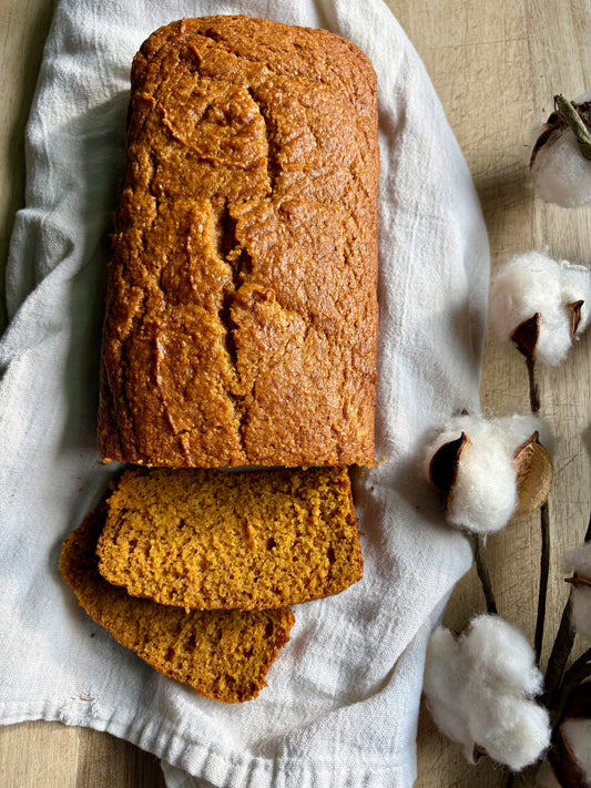 A loaf of golden-brown pumpkin bread with two slices cut and placed in front, resting on a white linen cloth with cotton bolls on a light wood surface.
