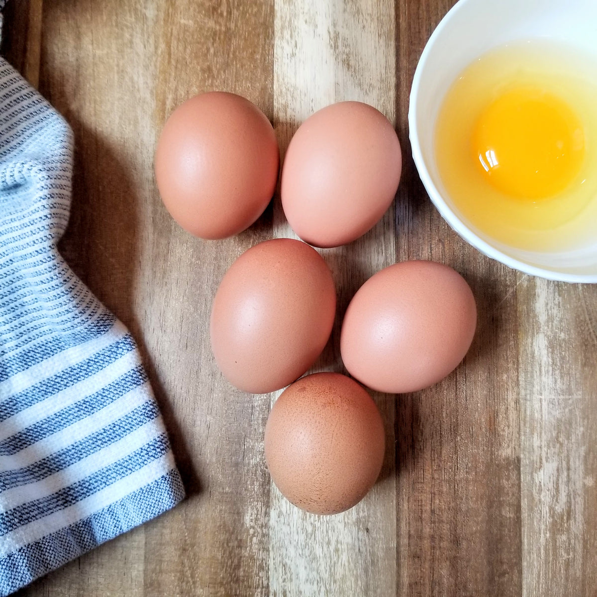 5 eggs on a wood cutting board with one cracked egg in a bowl beside a kitchen towel